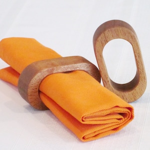 May include: An orange folded napkin is held by a wooden napkin ring. A second wooden napkin ring is next to the napkin. The napkin rings are oval-shaped and made of light brown wood. The napkin is a bright orange color.