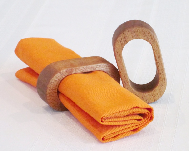 May include: An orange folded napkin is held by a wooden napkin ring. A second wooden napkin ring is next to the napkin. The napkin rings are oval-shaped and made of light brown wood. The napkin is a bright orange color.
