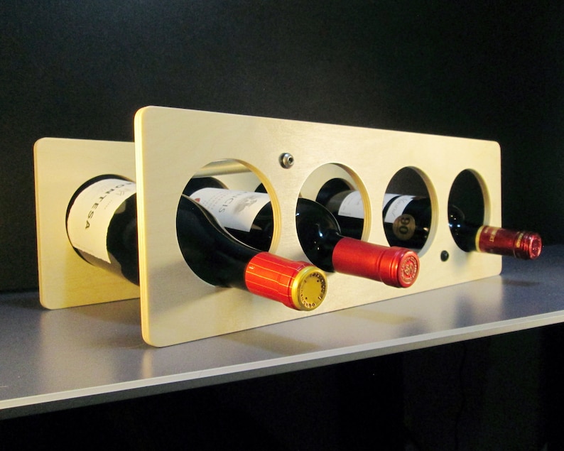 May include: A light-colored wooden wine rack with four circular openings, each holding a bottle of red wine. The bottles have red and gold foil around the neck. The rack is on a gray surface against a black background.