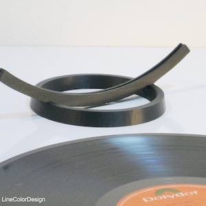 May include: A black vinyl record with an orange and white label is in the foreground. A black record holder with a curved design is in the center. The record holder is made of a dark gray material. The background is white.