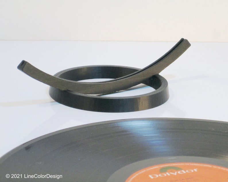 May include: A black vinyl record with an orange and white label is in the foreground. A black record holder with a curved design is in the center. The record holder is made of a dark gray material. The background is white.