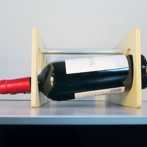 May include: A wooden wine rack with a bottle of red wine. The rack is made of light-colored wood and has a horizontal metal bar. The wine bottle is black with a white label and a red cap.
