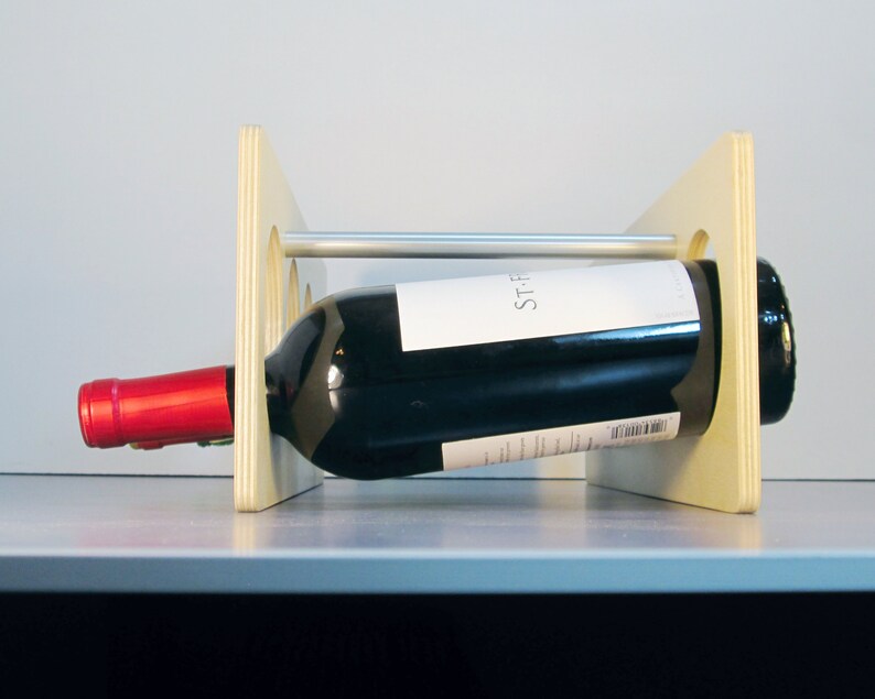 May include: A wooden wine rack with a bottle of red wine. The rack is made of light-colored wood and has a horizontal metal bar. The wine bottle is black with a white label and a red cap.