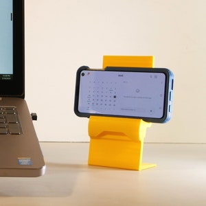 K&ouml;nnte beinhalten: Ein leuchtend gelber Telefonst&auml;nder, der ein Smartphone mit blauem Geh&auml;use h&auml;lt. Das Telefon zeigt einen Kalender an. Im Hintergrund ist ein Laptop zu sehen.