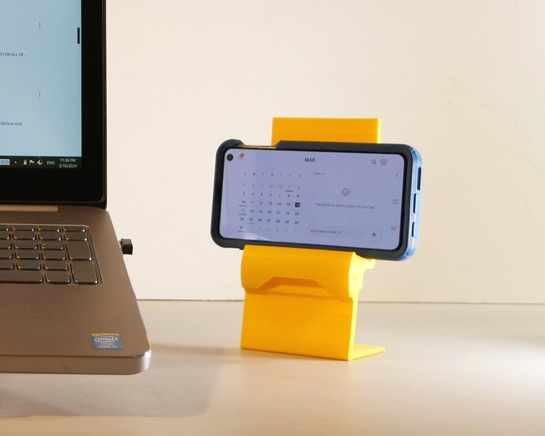 K&ouml;nnte beinhalten: Ein leuchtend gelber Telefonst&auml;nder, der ein Smartphone mit blauem Geh&auml;use h&auml;lt. Das Telefon zeigt einen Kalender an. Im Hintergrund ist ein Laptop zu sehen.