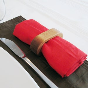 May include: A red cloth napkin is held by a wooden napkin ring. The napkin is placed on a brown placemat, with a silver knife to the side. The background is a white tablecloth. This image is suitable for a dining or table setting.