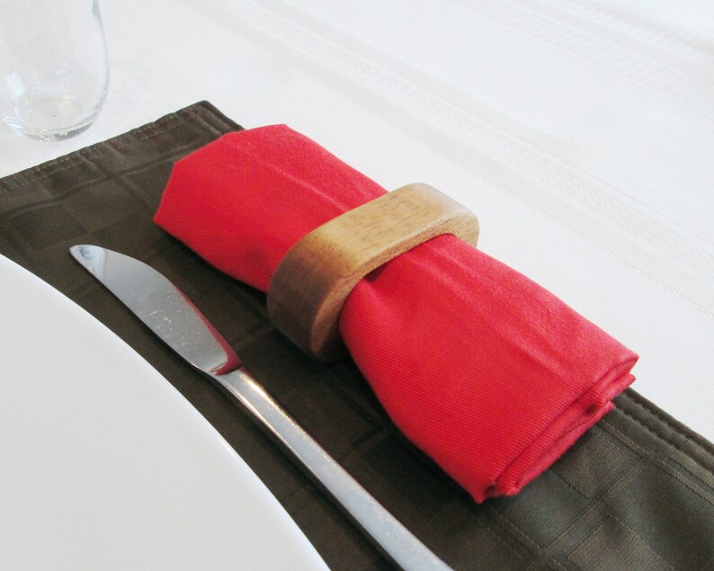 May include: A red cloth napkin is held by a wooden napkin ring. The napkin is placed on a brown placemat, with a silver knife to the side. The background is a white tablecloth. This image is suitable for a dining or table setting.