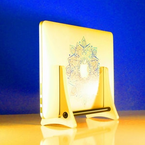 May include: A white laptop with a mandala design in blue, green, and pink, resting on a white wooden stand. The laptop is angled, revealing the floral pattern. The stand has a black support bar. The laptop is likely around 30 cm in length.