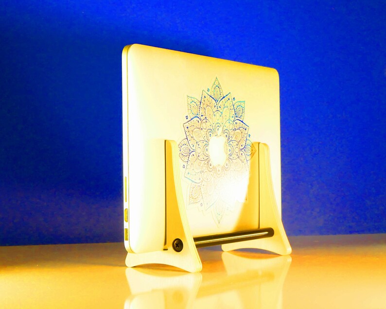 May include: A white laptop with a mandala design in blue, green, and pink, resting on a white wooden stand. The laptop is angled, revealing the floral pattern. The stand has a black support bar. The laptop is likely around 30 cm in length.