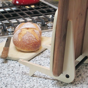 May include: A wooden breadboard and knife rest with a light wood frame, holding a wooden cutting board. A loaf of bread and a bread knife rest on a wooden cutting board. The kitchen setting includes a gas stove and a granite countertop.