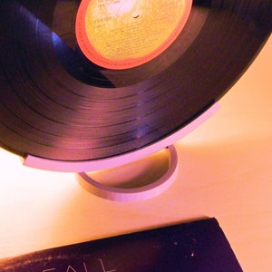 May include: A black vinyl record with a red and yellow label is displayed on a white stand. The record is partially visible, showcasing its grooves. A second album cover is visible in the foreground. The setup suggests a music or vintage audio theme.