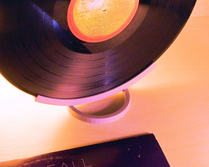 May include: A black vinyl record with a red and yellow label is displayed on a white stand. The record is partially visible, showcasing its grooves. A second album cover is visible in the foreground. The setup suggests a music or vintage audio theme.