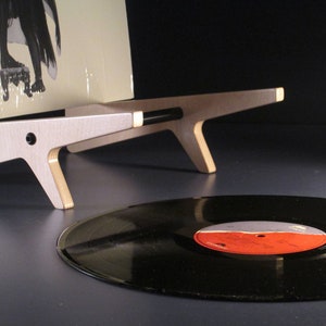 May include: A wooden record stand with a minimalist design, holding a vinyl record album. The stand is light-colored with a unique angular shape. A black vinyl record is in the foreground.