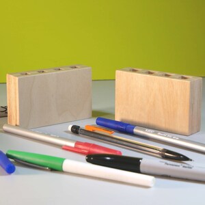 May include: Two unfinished wooden pen holders with multiple holes for pens and pencils. Assorted pens and markers in blue, green, red, black, and orange are scattered around the holders.