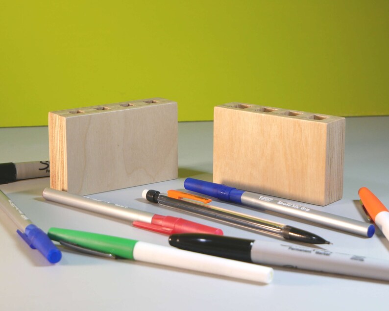 May include: Two unfinished wooden pen holders with multiple holes for pens and pencils. Assorted pens and markers in blue, green, red, black, and orange are scattered around the holders.