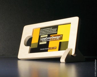 May include: A wooden business card holder with a rectangular frame and a supporting base. The holder displays a business card with the LineColorDesign logo and website information. The design is minimalist and modern, with a natural wood finish.
