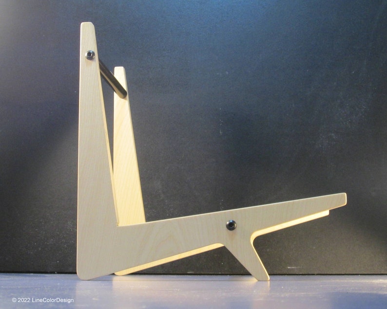 May include: A light-colored wooden guitar stand with a minimalist design. The stand features a triangular frame with a horizontal support and a black metal bar. The stand is held together with black screws. The design is modern and functional.