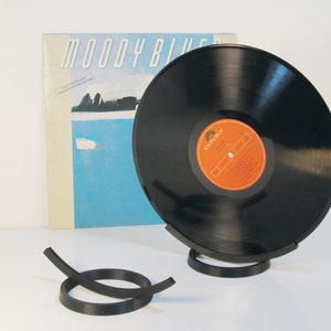 May include: A black vinyl record displayed in a black metal stand, with the album cover of "Moody Blues" in the background. The record has an orange and white label. A separate black metal record holder is in the foreground.