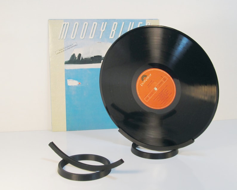 May include: A black vinyl record displayed in a black metal stand, with the album cover of "Moody Blues" in the background. The record has an orange and white label. A separate black metal record holder is in the foreground.