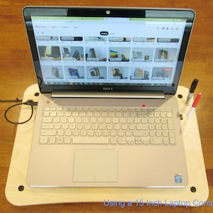 May include: A silver laptop computer on a light wood-colored laptop desk. The laptop is open, displaying a website with multiple images. A black and red marker are in a holder on the desk. The text at the bottom says "Using a 15 inch Laptop Computer."