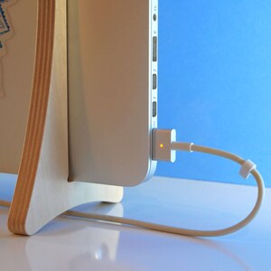 May include: A laptop computer connected to a power adapter via a beige cable. The laptop is mounted on a light-coloured wooden stand with a blue and white design. The background is a gradient of blue.
