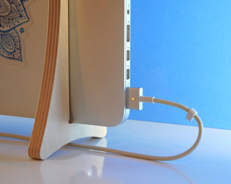 May include: A laptop computer connected to a power adapter via a beige cable. The laptop is mounted on a light-coloured wooden stand with a blue and white design. The background is a gradient of blue.