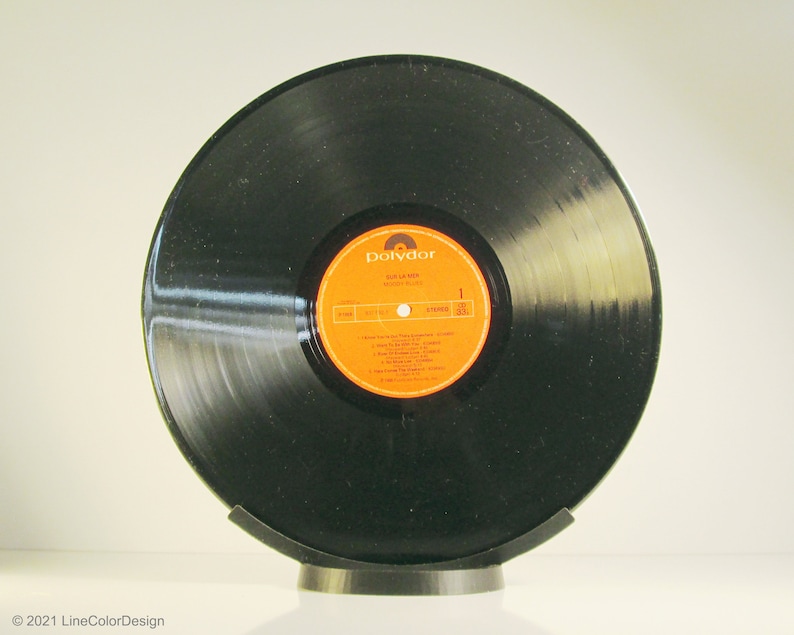 May include: A black vinyl record with an orange and yellow label, displayed on a black stand. The label reads "Sail La Mer" and "Moody Blues". The record is a vintage music item.