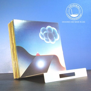 May include: A stack of art prints with a surreal landscape design featuring a blue sky, geometric shapes, and a white stand. The prints have a glossy finish and are displayed on a dark surface. The text "ORIGINAL LineColorDesign DESIGNED AND MADE IN USA" is visible.