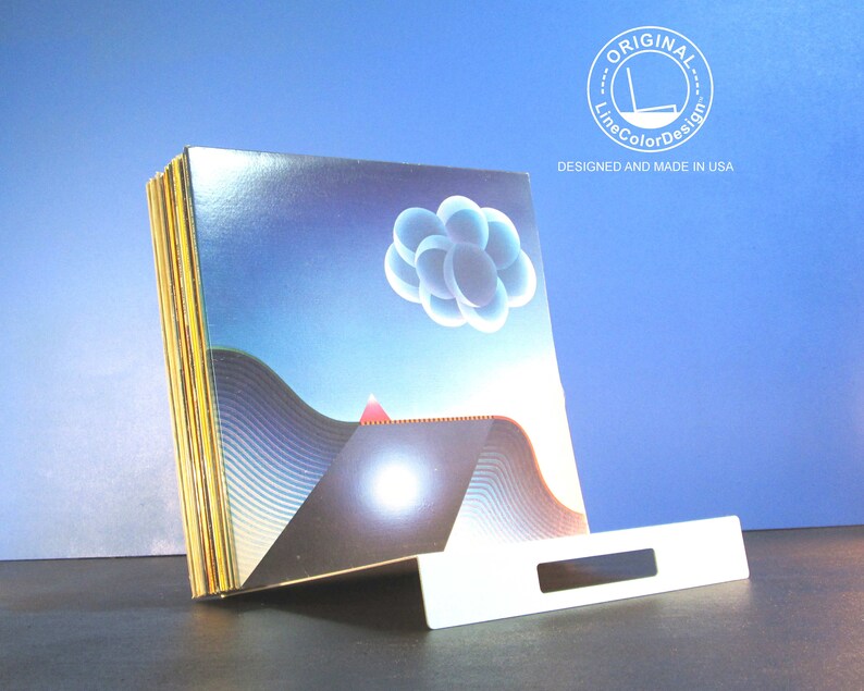 May include: A stack of art prints with a surreal landscape design featuring a blue sky, geometric shapes, and a white stand. The prints have a glossy finish and are displayed on a dark surface. The text "ORIGINAL LineColorDesign DESIGNED AND MADE IN USA" is visible.