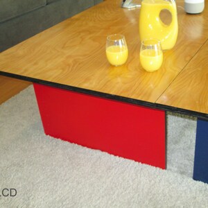 May include: A modern coffee table with a light wood grain top and red and blue supports. Two glasses of orange juice and a pitcher are on the table. The table is on a light gray rug.