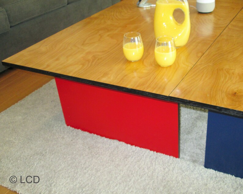 May include: A modern coffee table with a light wood grain top and red and blue supports. Two glasses of orange juice and a pitcher are on the table. The table is on a light gray rug.