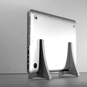 May include: A silver laptop is placed on a light grey wooden stand. The laptop's back is visible, highlighting its design. The stand has a minimalist design, offering support and elevating the computer. The stand is made of wood.