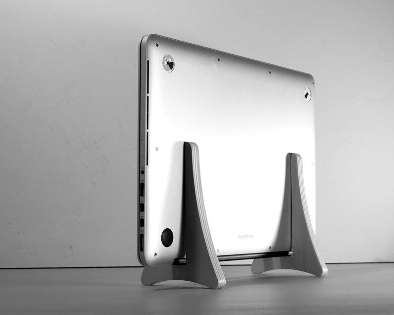 May include: A silver laptop is placed on a light grey wooden stand. The laptop's back is visible, highlighting its design. The stand has a minimalist design, offering support and elevating the computer. The stand is made of wood.