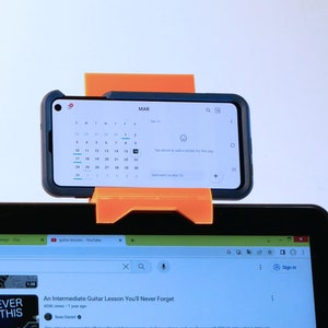 K&ouml;nnte beinhalten: Ein orangefarbener, 3D-gedruckter Telefonhalter mit einem Smartphone, das einen Kalender f&uuml;r M&auml;rz anzeigt. Das Telefon ist &uuml;ber einem Laptop-Bildschirm montiert, der ein YouTube-Video &uuml;ber Gitarrenunterricht zeigt. Der Halter ist so konzipiert, dass er das Telefon an Ort und Stelle h&auml;lt.
