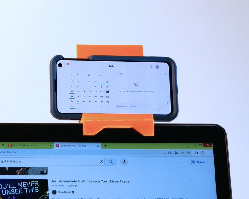 K&ouml;nnte beinhalten: Ein orangefarbener, 3D-gedruckter Telefonhalter mit einem Smartphone, das einen Kalender f&uuml;r M&auml;rz anzeigt. Das Telefon ist &uuml;ber einem Laptop-Bildschirm montiert, der ein YouTube-Video &uuml;ber Gitarrenunterricht zeigt. Der Halter ist so konzipiert, dass er das Telefon an Ort und Stelle h&auml;lt.