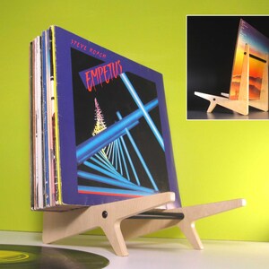 May include: A wooden record holder with a stack of vinyl records. The record holder is made of light-colored wood and has a unique, angular design. The album cover visible features the title "Empetus" and a geometric design in blue and red.