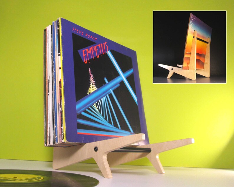 May include: A wooden record holder with a stack of vinyl records. The record holder is made of light-colored wood and has a unique, angular design. The album cover visible features the title "Empetus" and a geometric design in blue and red.