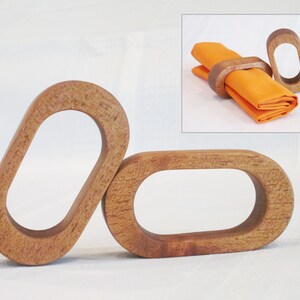 May include: Three wooden napkin rings with an oval design. The rings are made of light brown wood and have a smooth, polished finish. An inset image shows a ring holding an orange napkin.