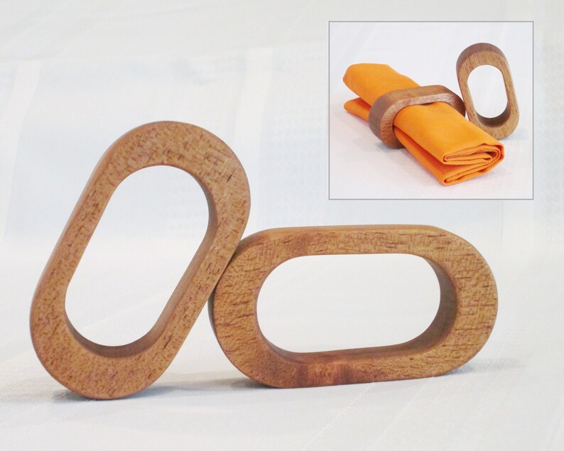 May include: Three wooden napkin rings with an oval design. The rings are made of light brown wood and have a smooth, polished finish. An inset image shows a ring holding an orange napkin.