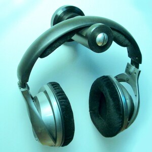 May include: Silver and black over-ear headphones with large, round earcups and a padded headband. The headphones are mounted on a black wall-mounted holder. The earcups have a soft, velvety texture.