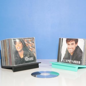 May include: Two CD holders, one black and one teal, display stacks of CDs. The black holder displays the "Bodyguard" soundtrack. The teal holder displays a CD by Cody Webb. A single CD sits on the white surface.