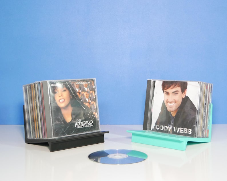 May include: Two CD holders, one black and one teal, display stacks of CDs. The black holder displays the "Bodyguard" soundtrack. The teal holder displays a CD by Cody Webb. A single CD sits on the white surface.