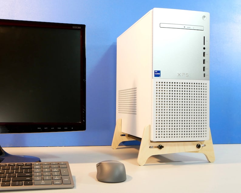 May include: A white and silver XPS computer tower with the Intel Core logo, set on a light wood stand. A black monitor, keyboard, and gray mouse are in the foreground. The background is a gradient of blue.