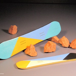 May include: Two snowboards with geometric designs in blue, yellow, and teal. One board is positioned at an angle, while the other lies flat. Small orange rocks are scattered around the boards. The boards have a wooden appearance.