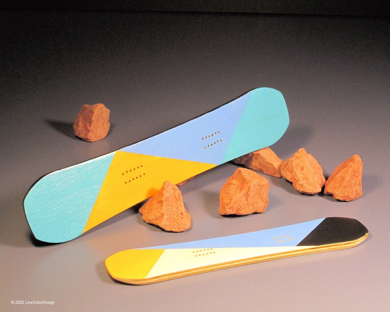 May include: Two snowboards with geometric designs in blue, yellow, and teal. One board is positioned at an angle, while the other lies flat. Small orange rocks are scattered around the boards. The boards have a wooden appearance.