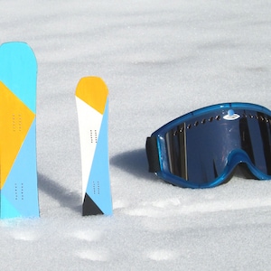 May include: Two snowboards with geometric designs in blue, yellow, and white, and a pair of blue ski goggles with a reflective lens, all resting on a snowy surface. The snowboards have a series of small holes along the center.