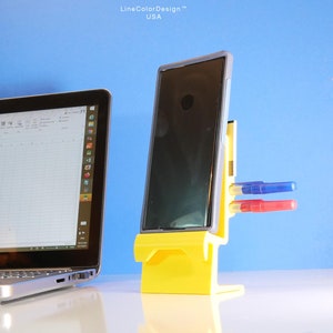 May include: A yellow smartphone stand with a black smartphone and two pens. The stand has a slot for the phone and two holes for pens. A laptop is visible in the background. The text "LineColorDesign USA" is in the top right corner.