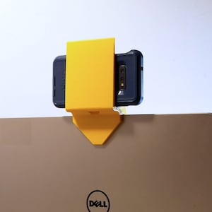 K&ouml;nnte beinhalten: Ein gelber, 3D-gedruckter Telefonhalter, der an einem Laptop befestigt ist. Das Telefon befindet sich in einer dunkelblauen H&uuml;lle. Der Laptop ist ein Dell-Modell mit silbernem Finish. Der Telefonhalter ist so konzipiert, dass er am Laptop-Bildschirm befestigt werden kann.