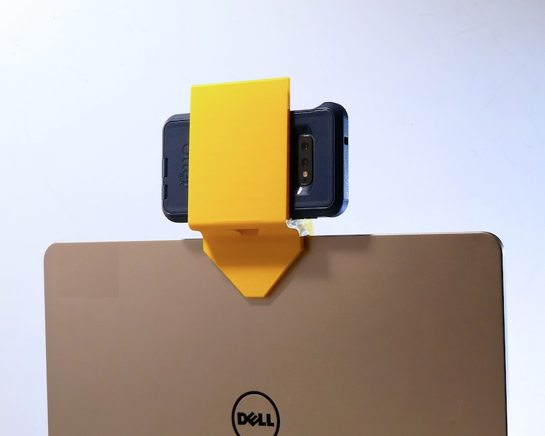 K&ouml;nnte beinhalten: Ein gelber, 3D-gedruckter Telefonhalter, der an einem Laptop befestigt ist. Das Telefon befindet sich in einer dunkelblauen H&uuml;lle. Der Laptop ist ein Dell-Modell mit silbernem Finish. Der Telefonhalter ist so konzipiert, dass er am Laptop-Bildschirm befestigt werden kann.