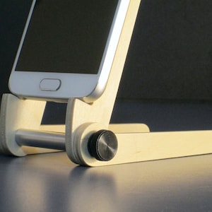 May include: A wooden phone stand with a silver metal bar and black knobs. A white smartphone is placed on the stand. The stand is made of light-colored wood and has a simple, functional design.
